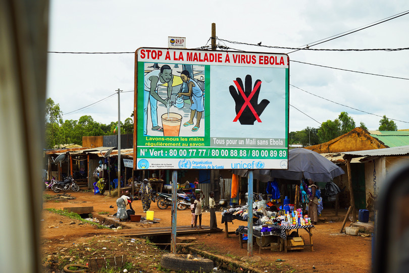 A wide shot of a dusty street market in Mali. A large, prominent billboard in the center displays a health poster about Ebola prevention. The poster is in French and features two main illustrations. On the left, two cartoon figures wash their hands with soap and water from a bucket. On the right, a large red 'X' crosses out an image of a hand, symbolizing a prohibition on handshakes. The text on the billboard reads 'STOP À LA MALADIE À VIRUS EBOLA !' (STOP THE EBOLA VIRUS DISEASE!), 'Lavons-nous les mains régulièrement au savon' (Let's wash our hands regularly with soap), and 'Tous pour un Mali sans Ebola' (All for an Ebola-free Mali).